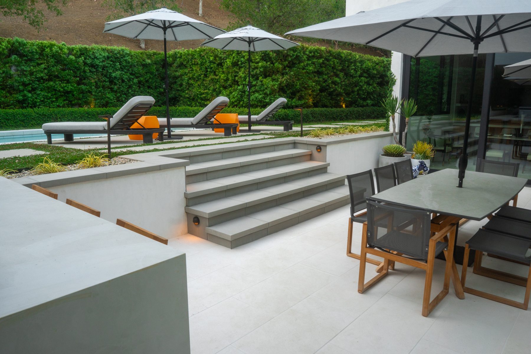 A multi-level modern patio with wide steps connecting a pool deck to a dining and lounge area, framed by market umbrellas and glass doors to the house