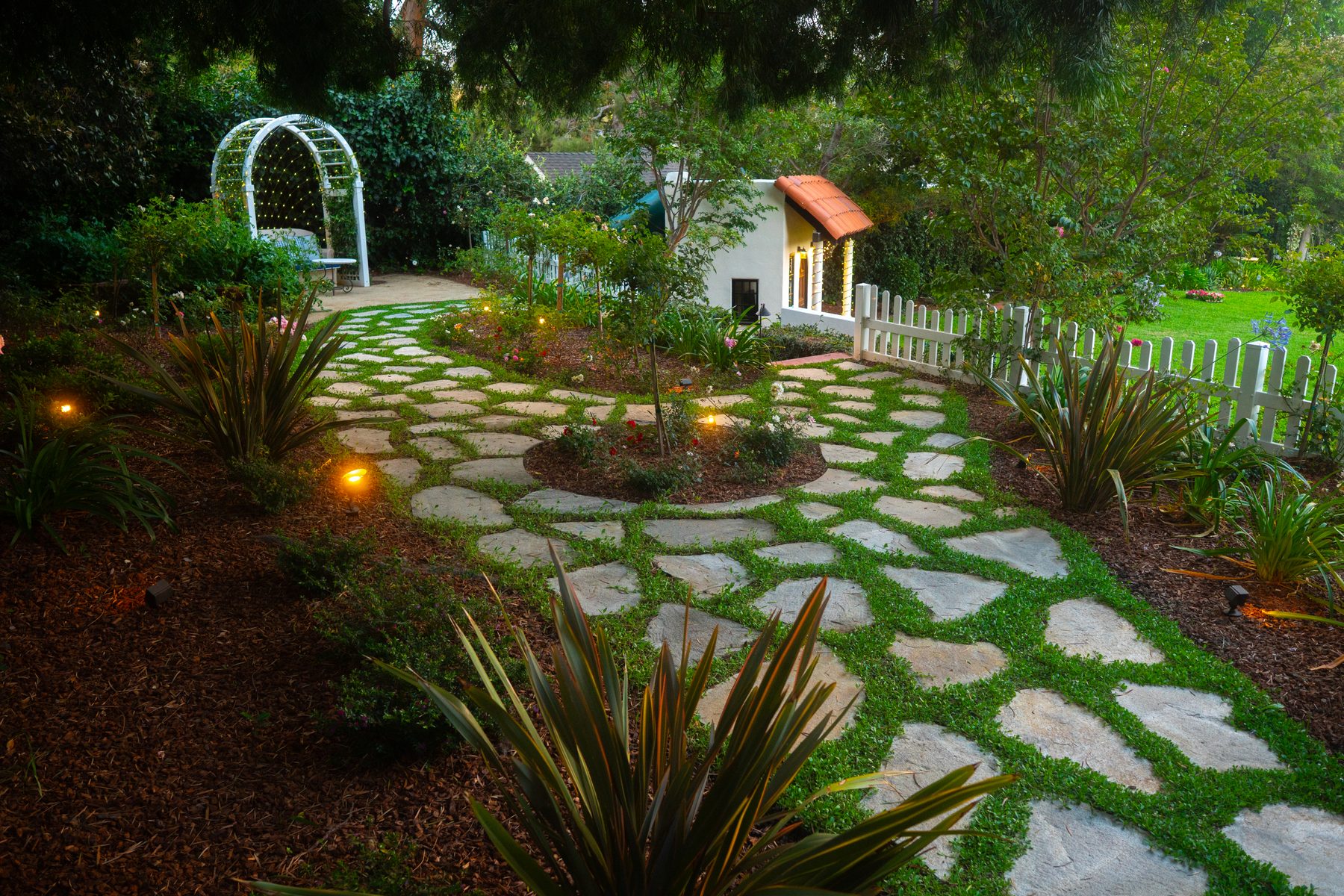 A flagstone garden path with grass-filled joints curving past an arbor toward a lit guest house at dusk