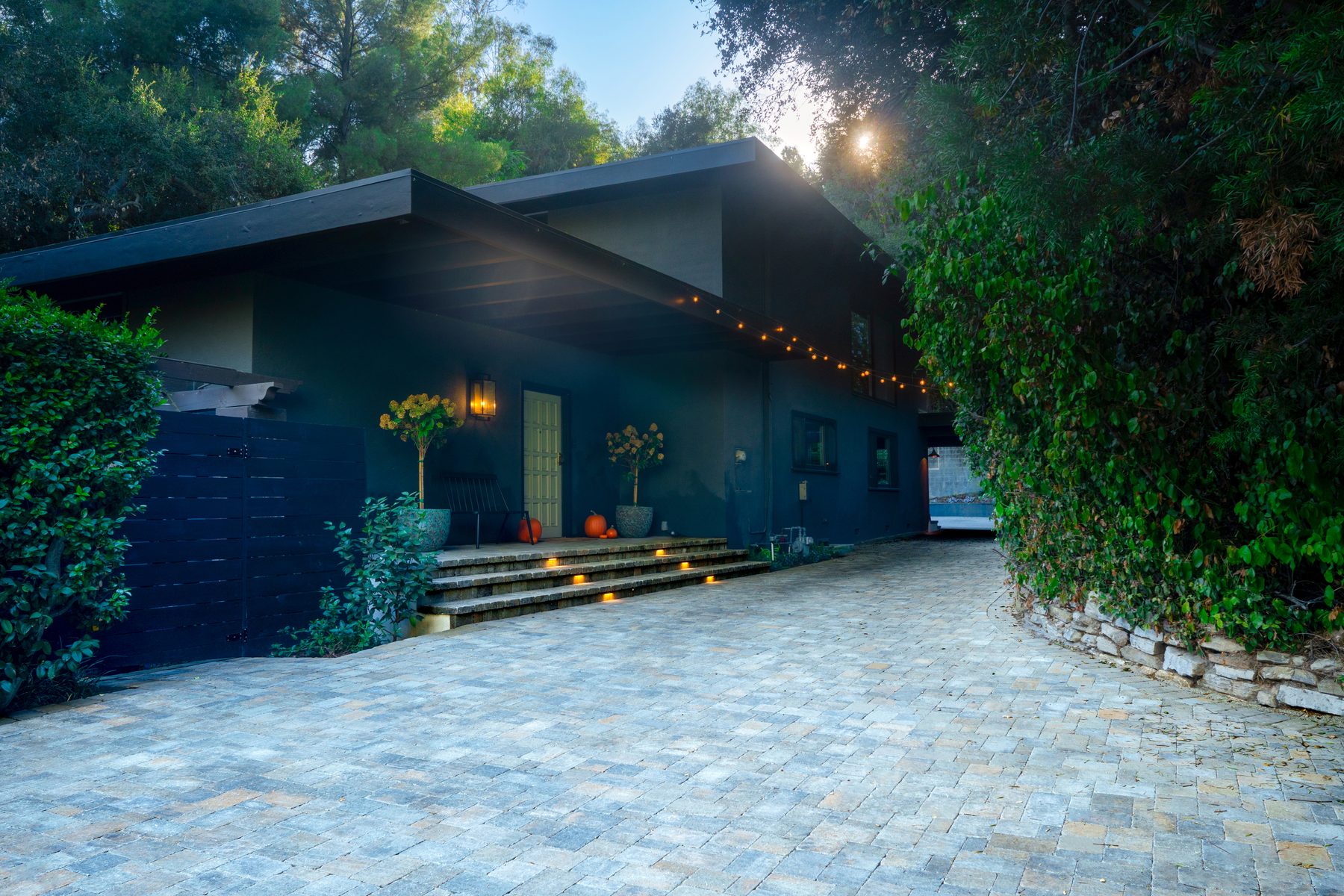 A paver driveway and entry courtyard at dusk in front of a dark modern home with string lights