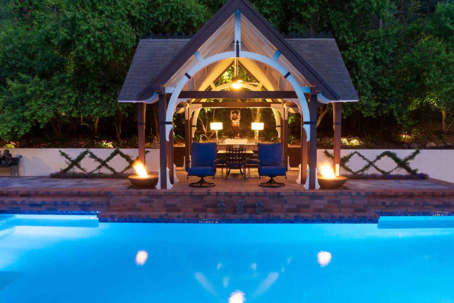 A timber-frame gazebo with white trellis braces over a poolside dining area at dusk, flanked by fire bowls