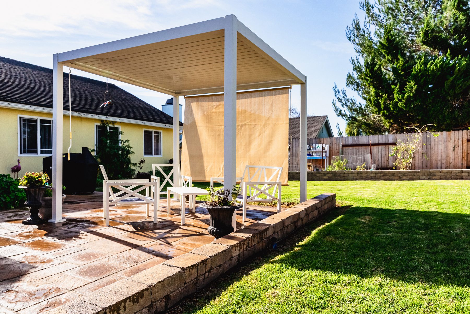A white aluminum pergola over a paver patio in Santa Maria, seen from the yellow stucco house with roses and a retaining wall in view