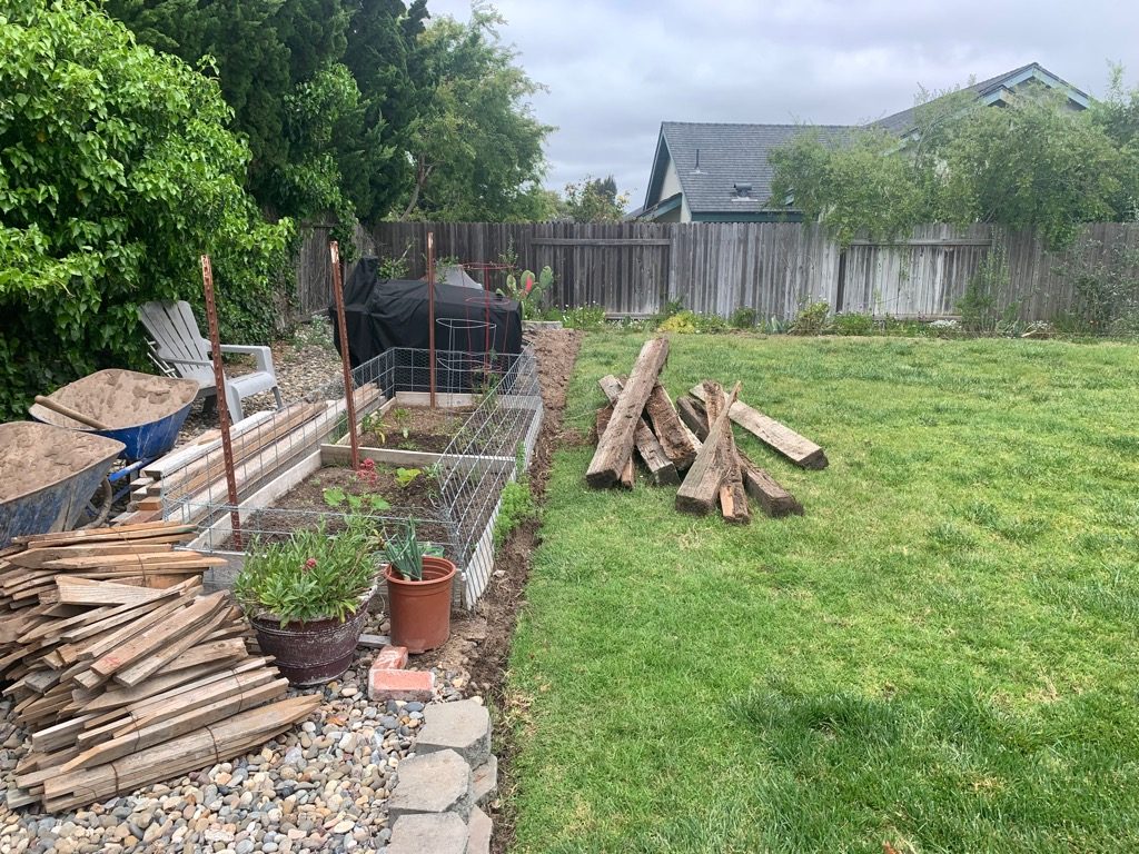 A bare Santa Maria backyard with raised garden beds, a wood pile, and a wheelbarrow of sand before construction began