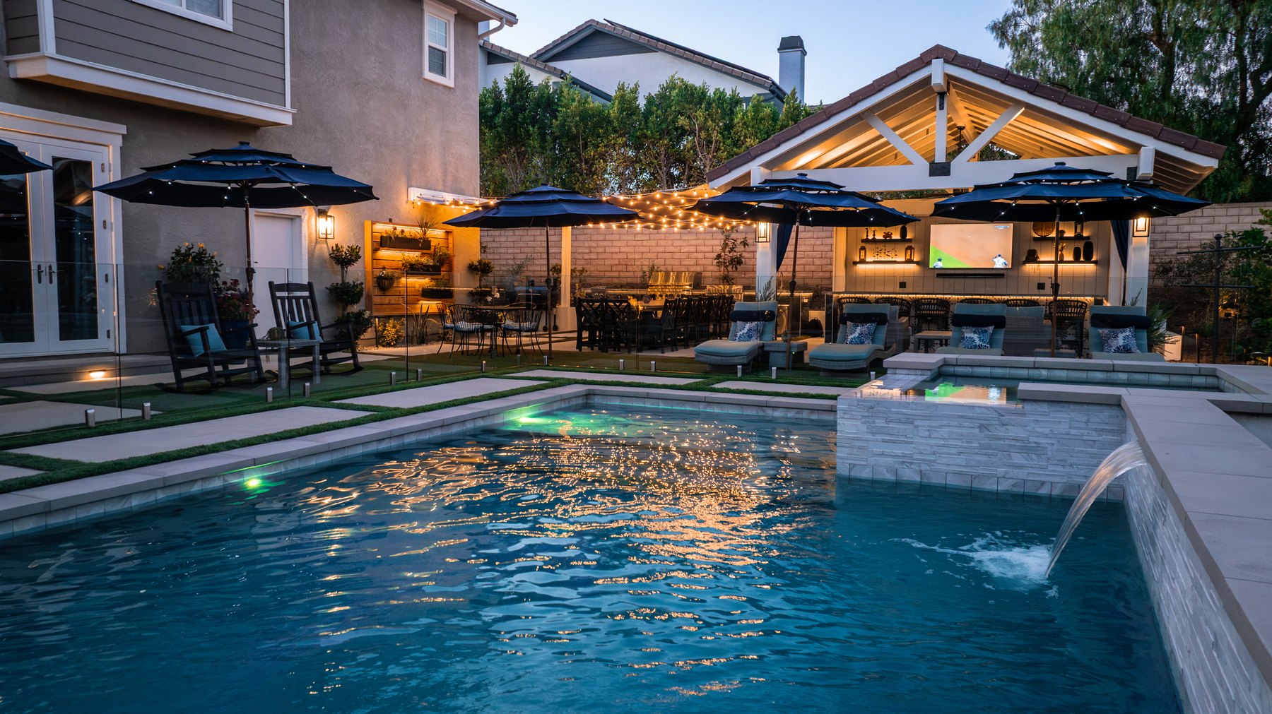 Evening backyard pool with a raised spa, pavilion, outdoor kitchen, string lights, and lounge seating