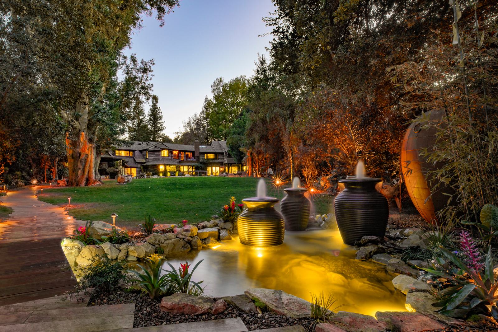 Three dark ribbed ceramic urn fountains spilling into a golden-lit pond at dusk, framed by a lawn, boardwalk, and mature trees