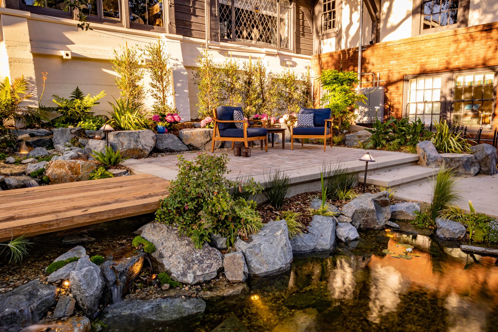 A wood-plank bridge crossing a lily-pad pond beside a paver patio with modern lounge chairs