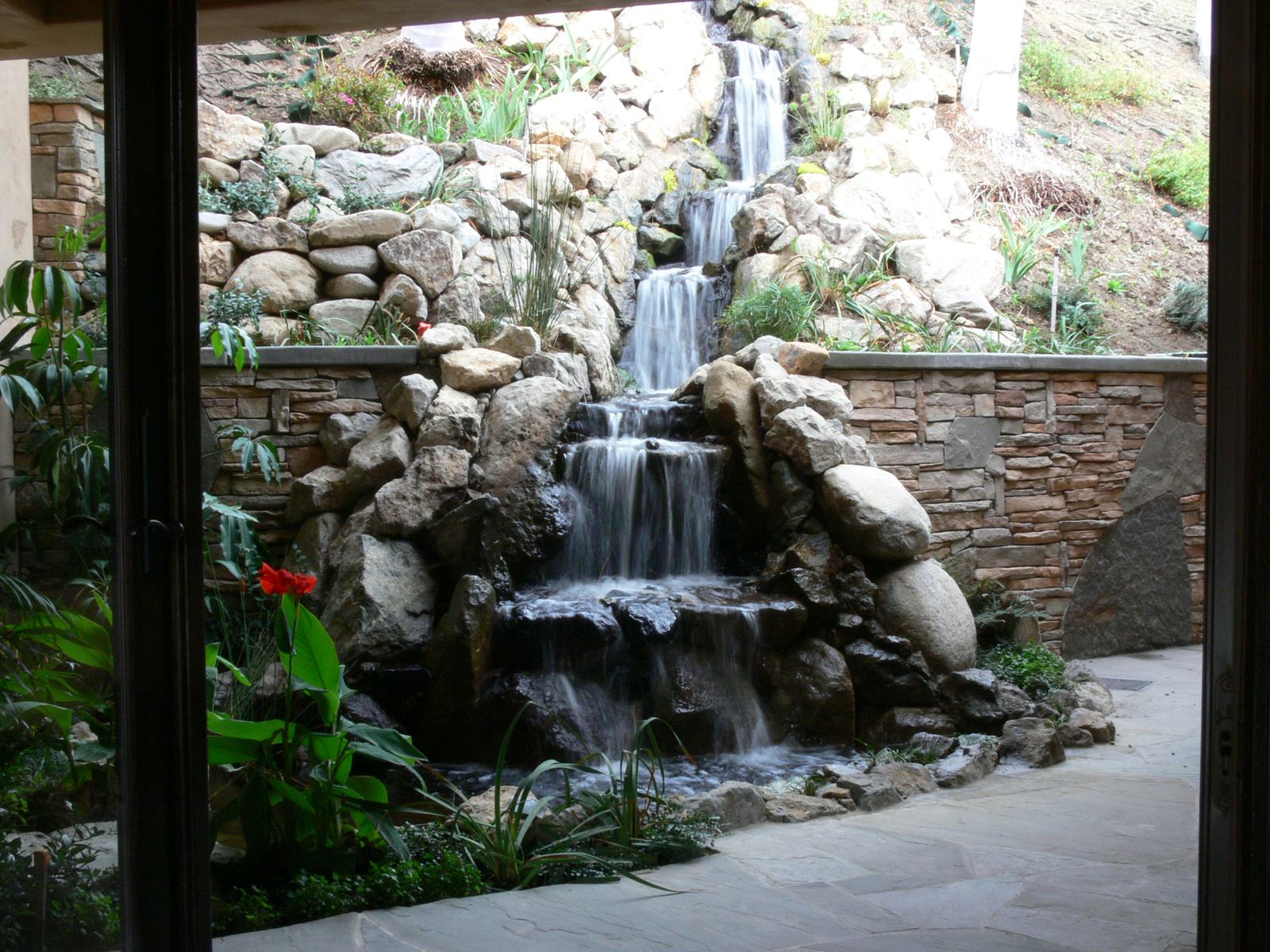 A multi-tier boulder waterfall cascading toward a pool, framed by a covered patio and stacked-stone retaining walls