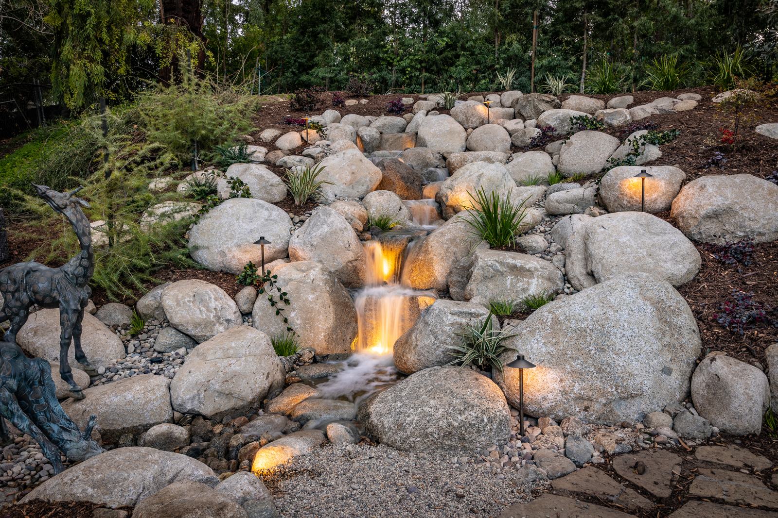 A tall multi-drop boulder waterfall lit at dusk, with bronze deer sculptures in the foreground garden