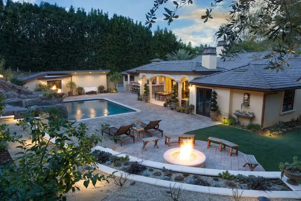 A large backyard boasts a lit fire-pit and a pool next to a rock formation.