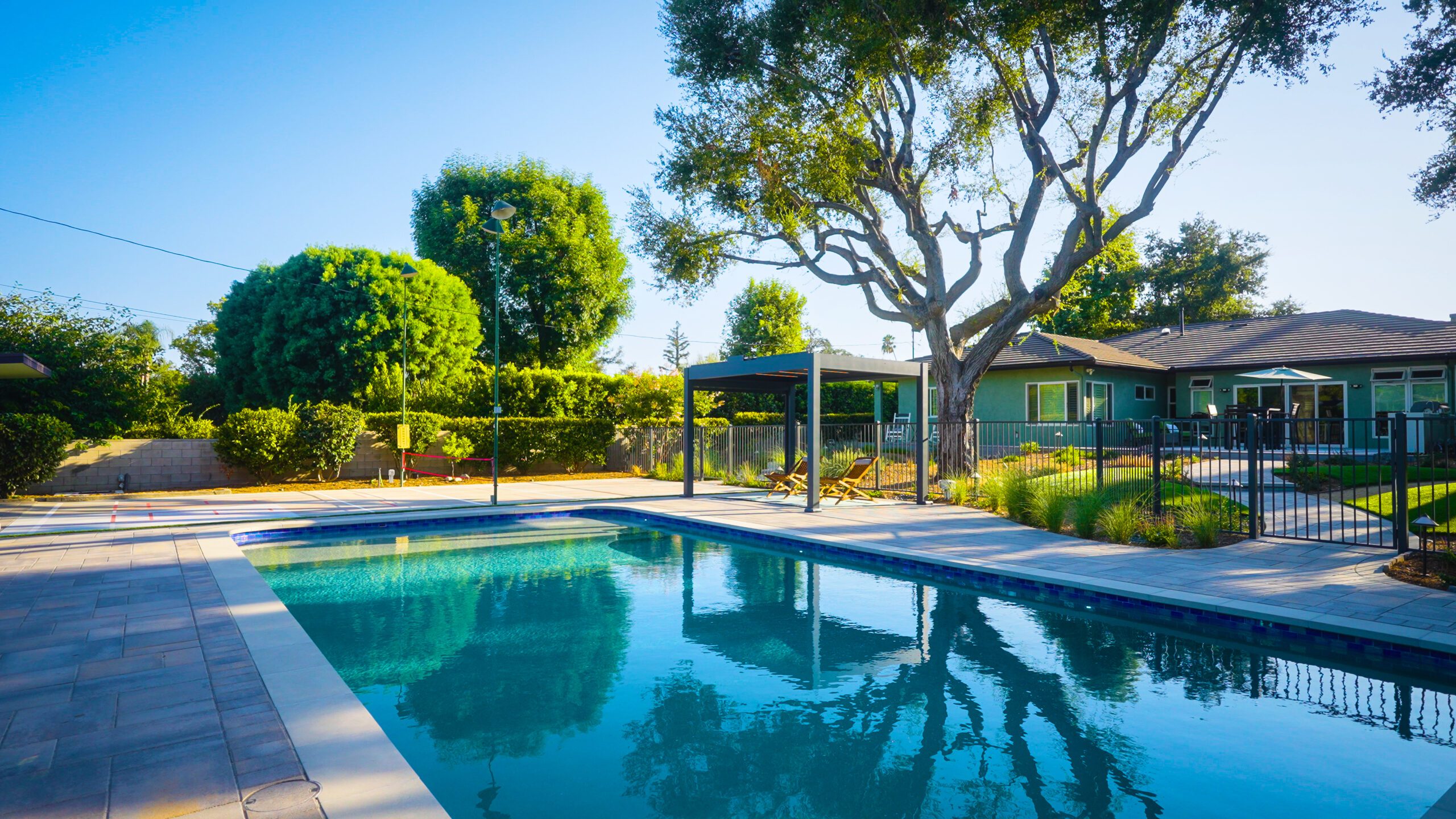 Wide view of contemporary backyard pool, pergola lounge, paver deck, fencing, and surrounding greenery creating a cohesive California outdoor living space.