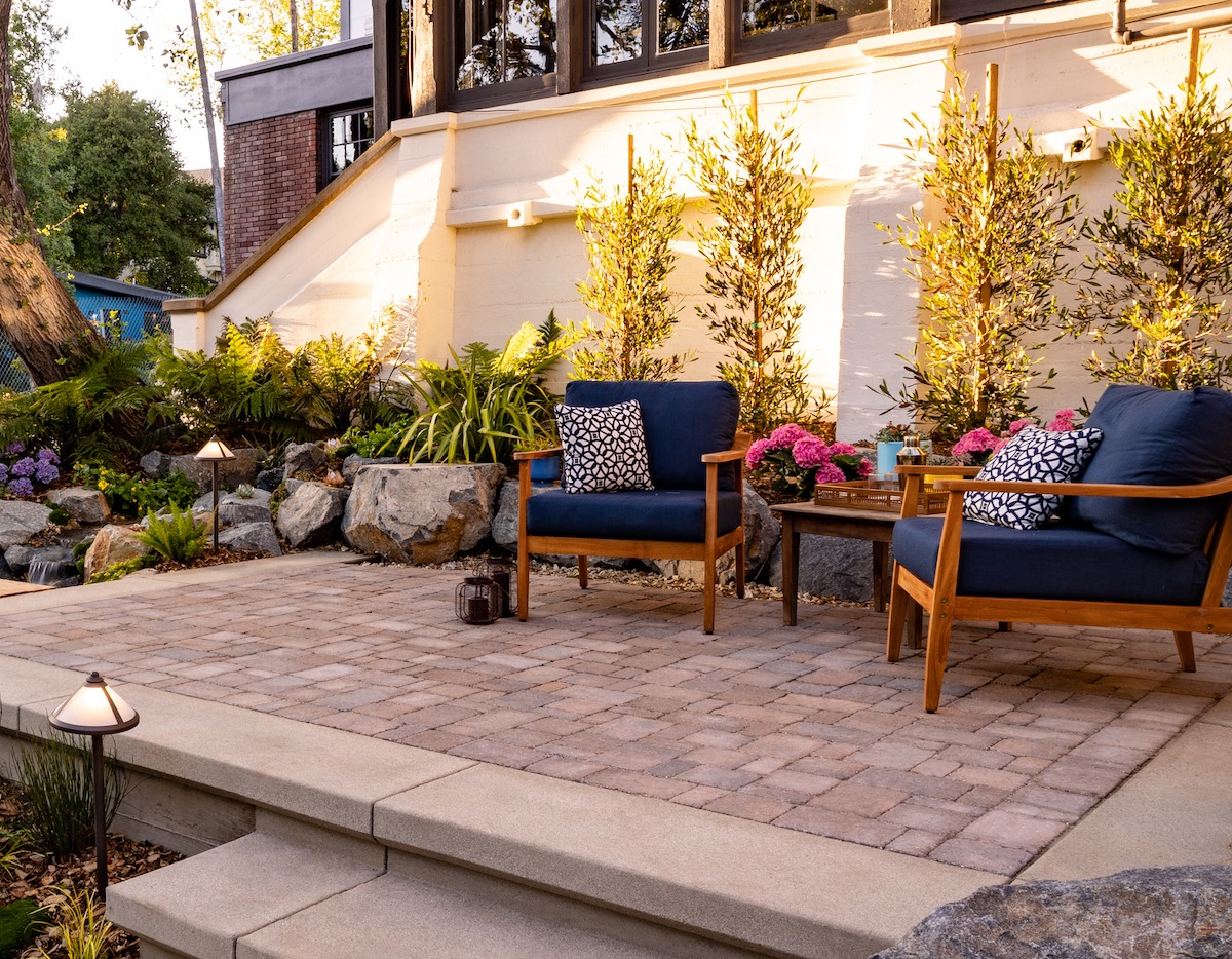 Paver patio with concrete steps, teak chairs, boulders, ferns, and olive trees lit by path lighting