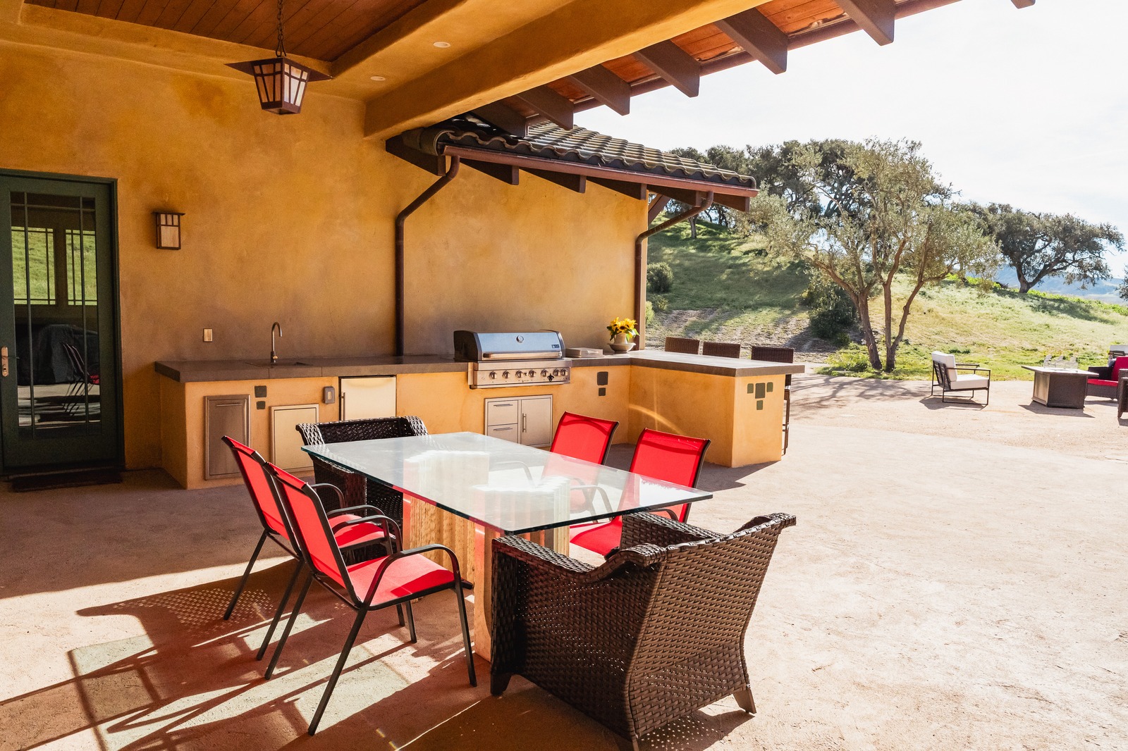 Covered patio outdoor kitchen with a stainless grill, sink, concrete counters, and dining table below oak-studded hills