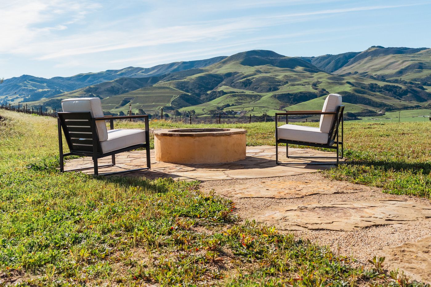 Flagstone circle with a stucco fire pit and two cushioned chairs on lawn below green rolling hills