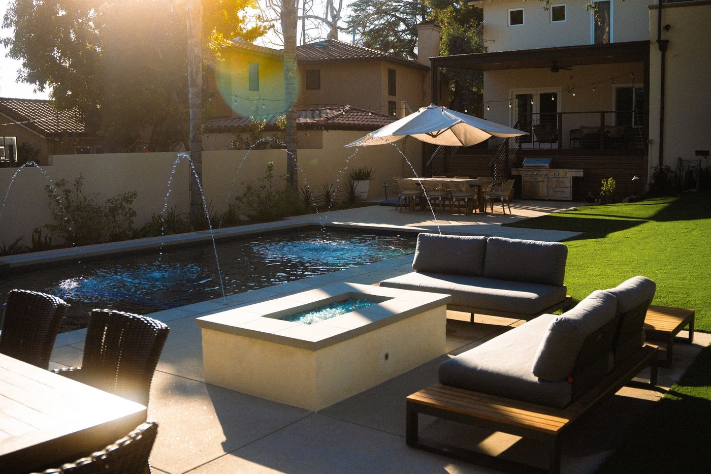 Backyard pool with arcing fountains, fire feature lounge, lawn, dining area, and outdoor kitchen in warm sunlight