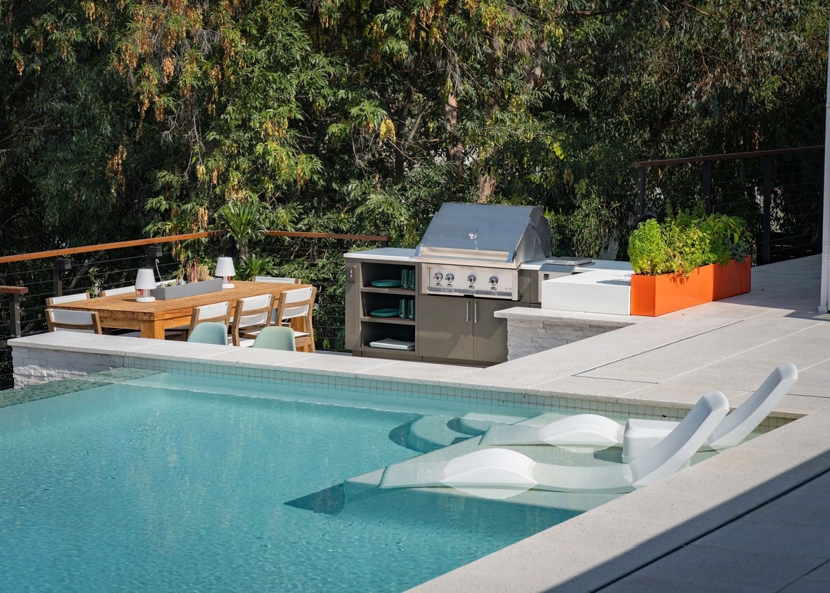 Pool with tanning-ledge loungers beside a stainless grill island with stone veneer and a wood dining table