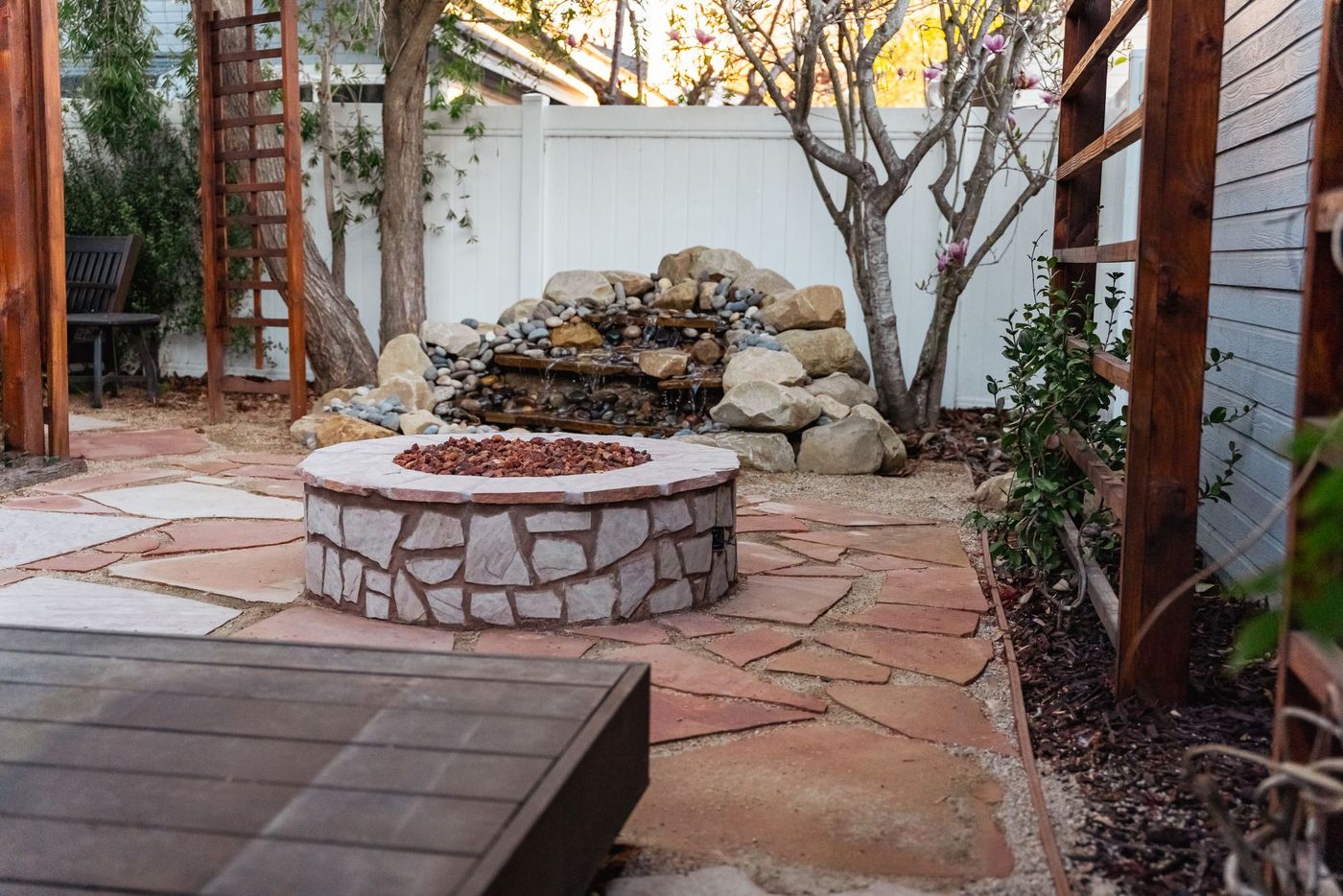 Custom backyard flagstone patio featuring a circular stone fire pit and natural rock waterfall water feature.