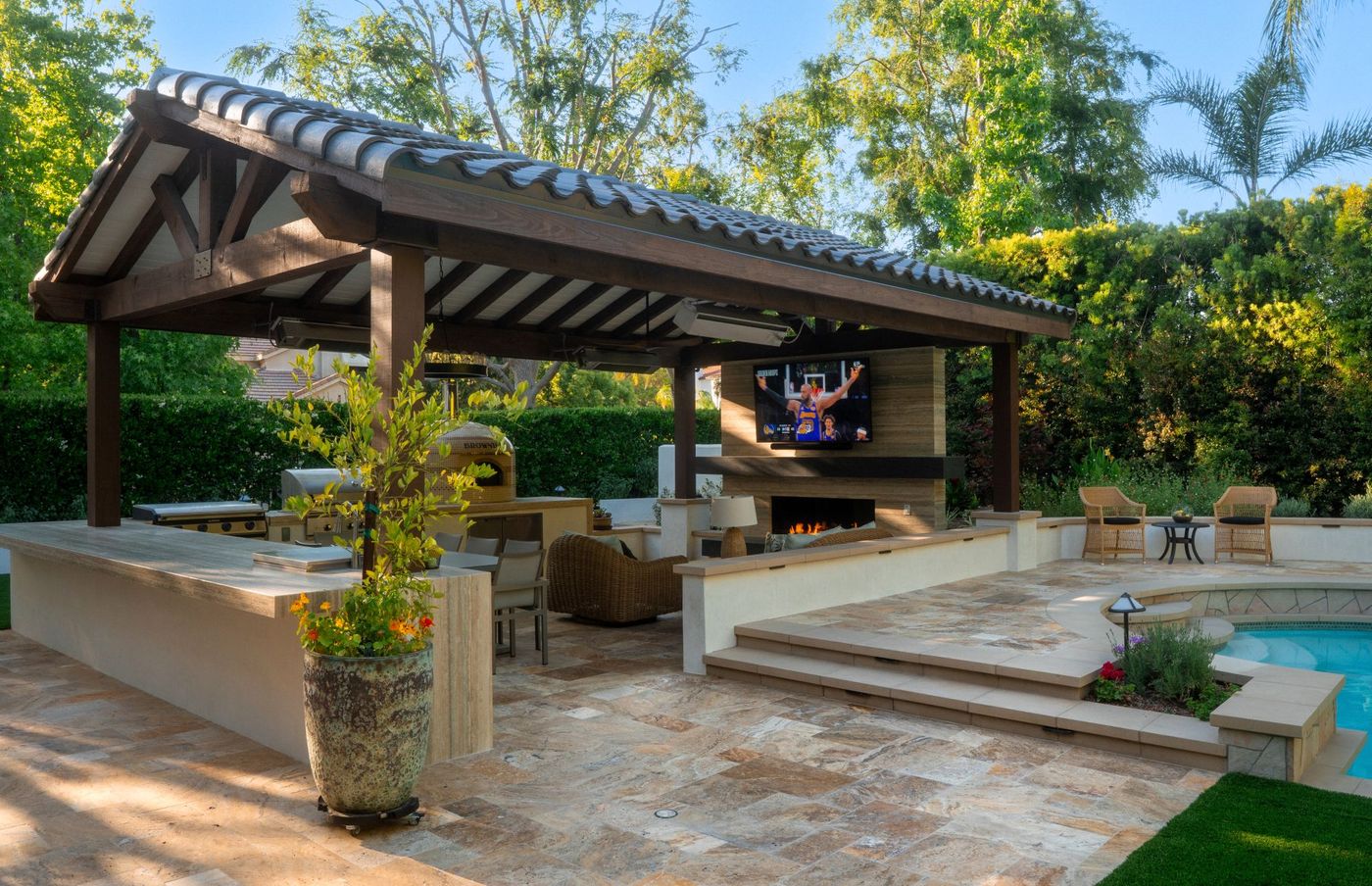 Covered outdoor kitchen and lounge with fireplace, TV, pool, and mature landscaping