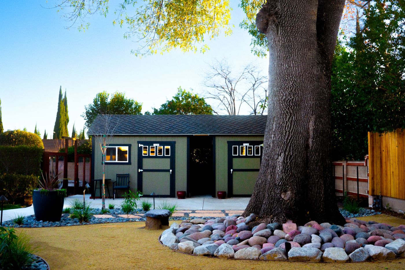 Backyard storage shed beneath an oak tree with river rock landscaping