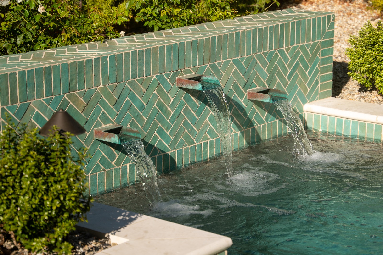 Copper scupper spouts pouring into a pool from a green herringbone-tile water feature wall