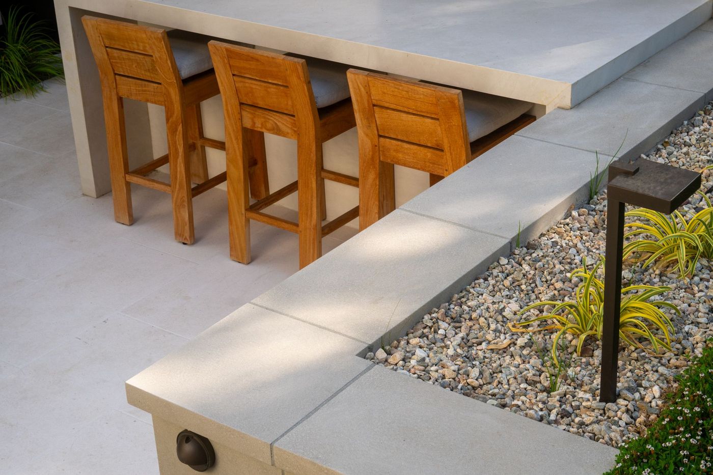 Wood bar stools tucked beneath a sleek concrete outdoor kitchen counter with decorative gravel landscape.