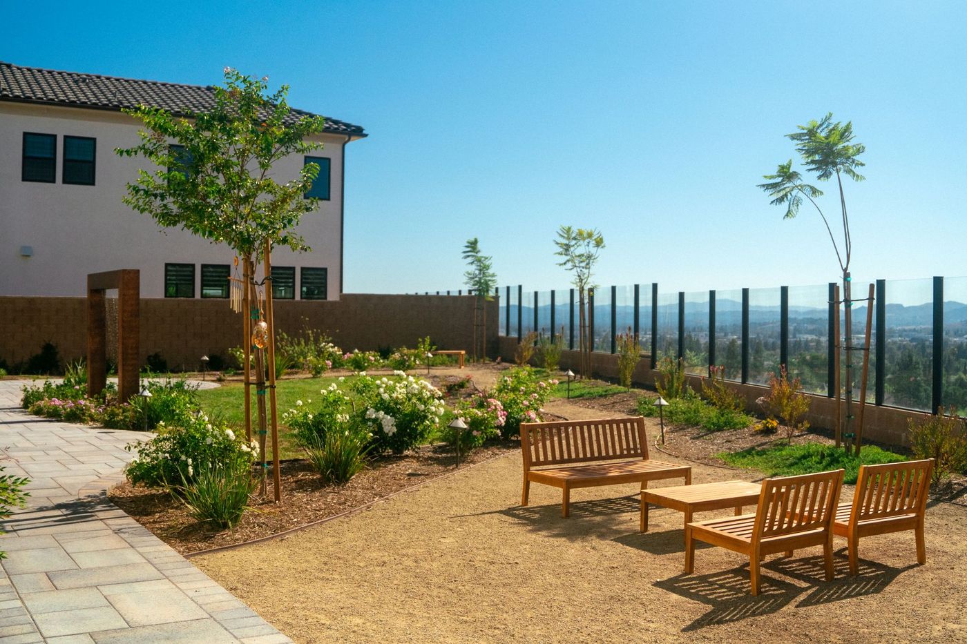 Wood bench seating placed along decomposed granite paths within a flowering courtyard garden bordered by glass fencing