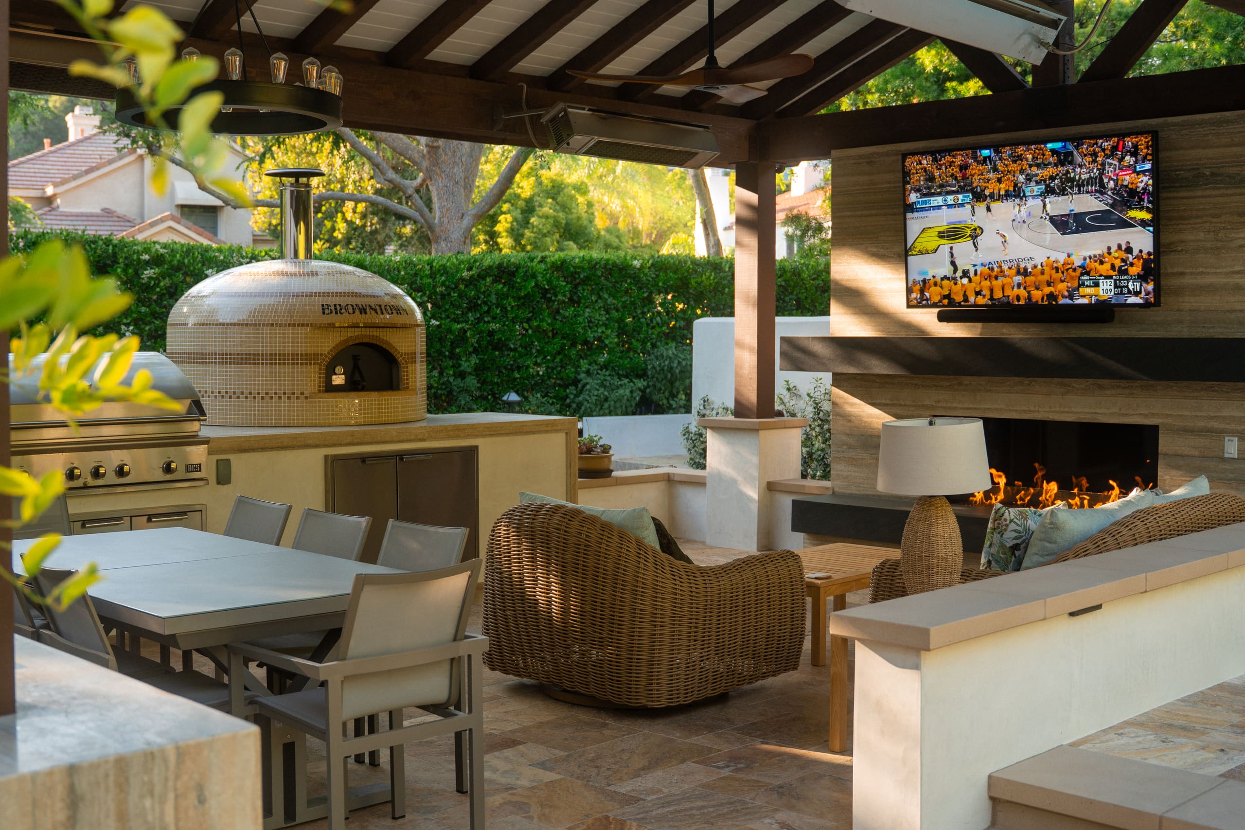 Covered outdoor living space featuring built-in grill, pizza oven, dining area, fireplace with mounted TV, and comfortable lounge seating in a landscaped backyard.