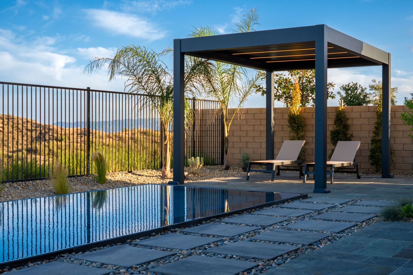 Wide view from beneath pergola showing outdoor dining, linear pool, stepping stone path, and expansive sky over contemporary backyard design.