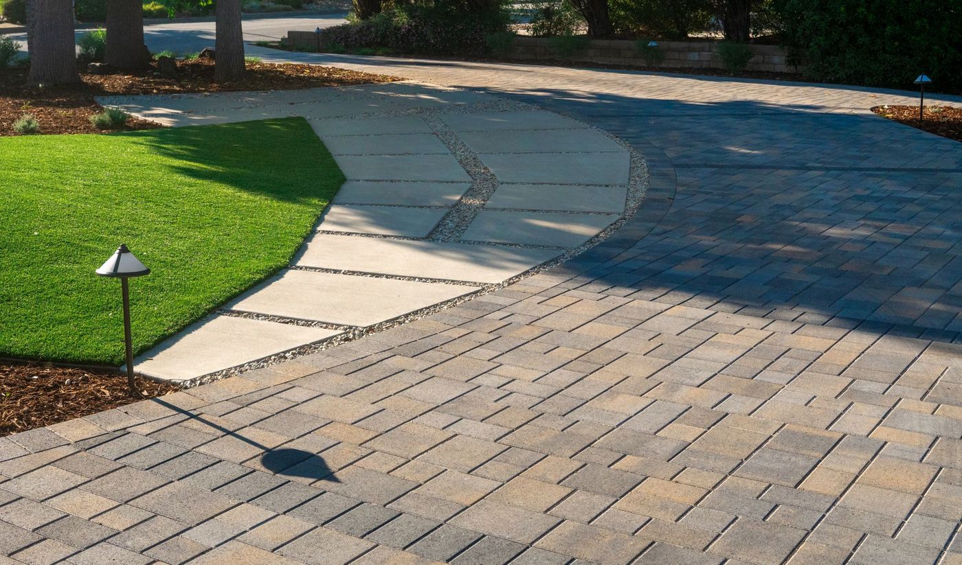 Smooth curved concrete walkway connecting lawn area to patterned paver driveway with integrated landscape lighting in a modern residential landscape.