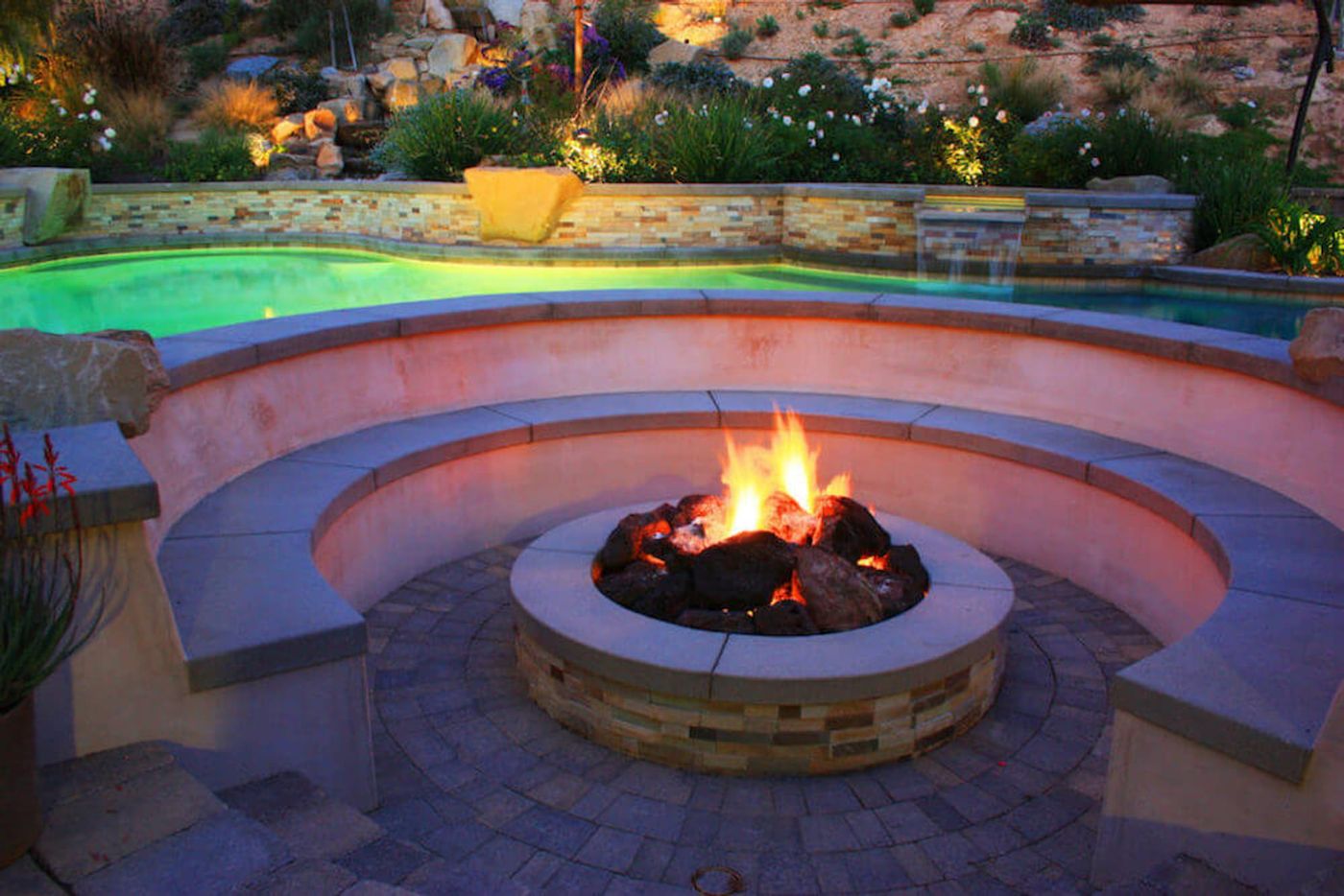 A circular fire pit ringed by a curved stone bench beside a lit pool at dusk
