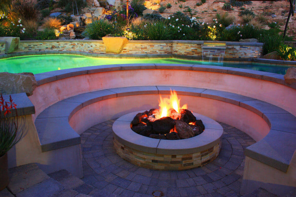Circular built-in fire pit with a full ring of curved stone benches, lit at dusk beside a pool