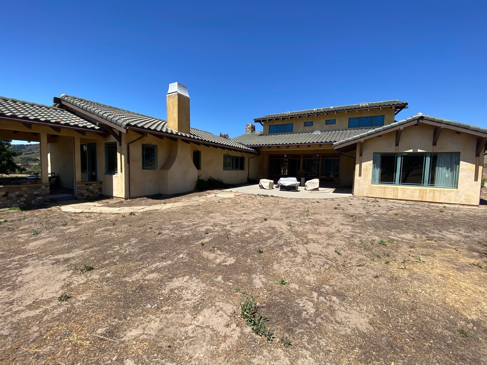 Before photo of the bare dirt yard behind a tile-roof stucco house with a small concrete patio