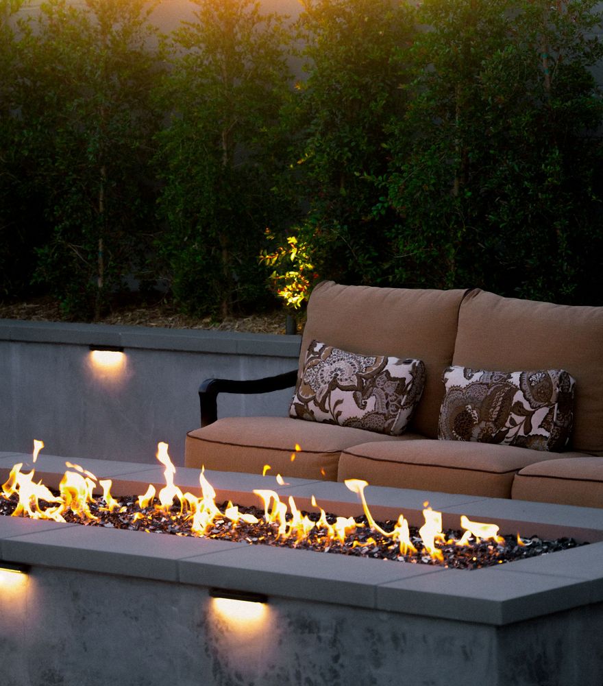 Modern linear fire pit with glass media in front of cushioned outdoor seating and landscape lighting.