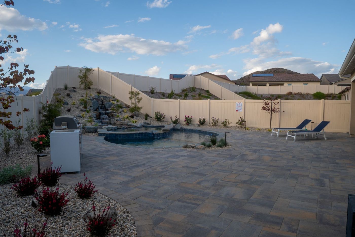 Custom backyard swimming pool with rock waterfall, large paver patio, and modern outdoor lounge area in a landscaped residential yard
