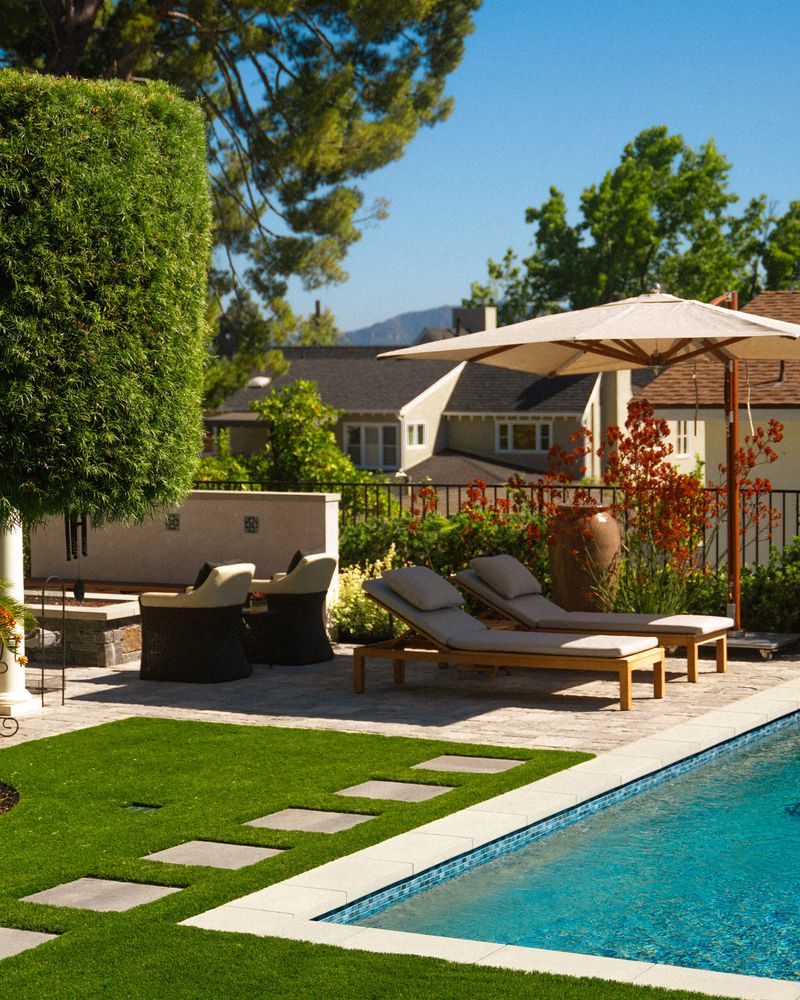 Poolside lounge chairs and patio umbrella beside a modern swimming pool in a professionally landscaped backyard.