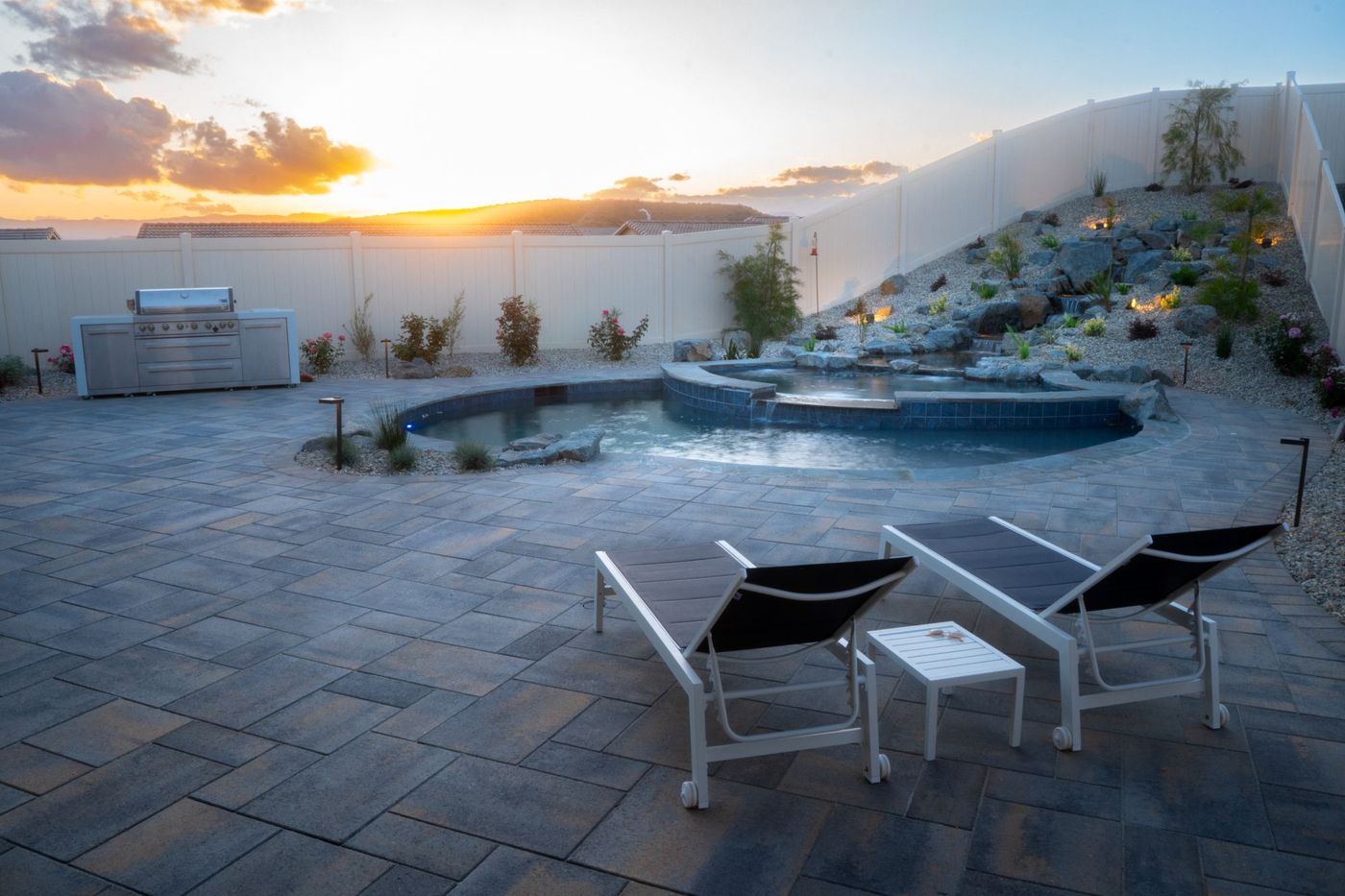 Luxury residential poolscape at sunset featuring freeform pool, illuminated rock waterfall, wide paver patio, and contemporary lounge chairs
