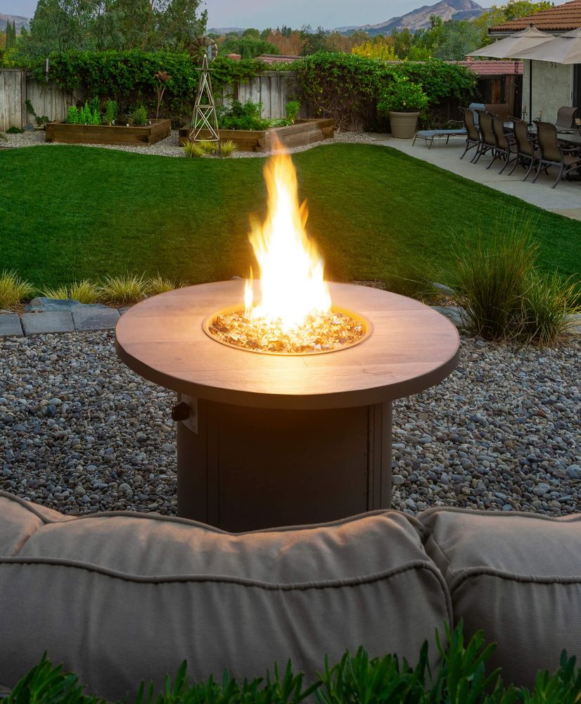 A round wood-topped gas fire table with a tall flame on a gravel patio beside a lawn and raised garden beds