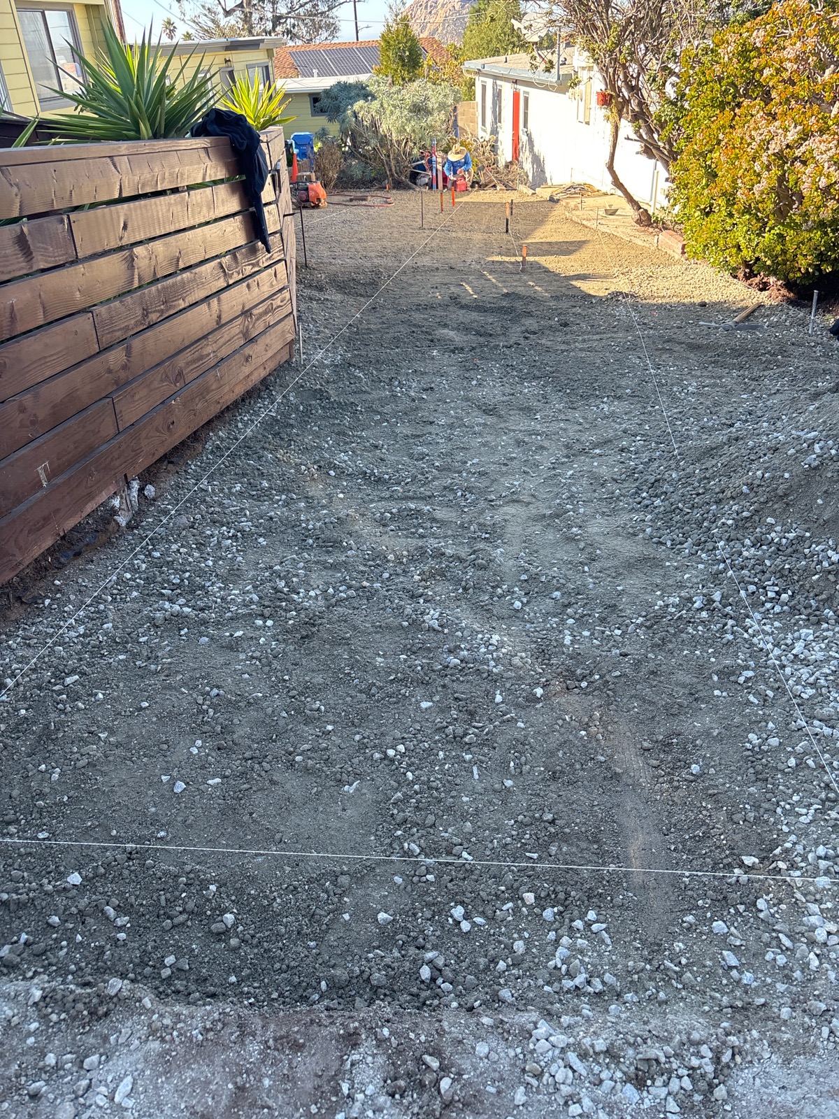 Graded gravel base marked out with string lines along a nearby Morro Bay driveway, with compaction equipment staged in the background