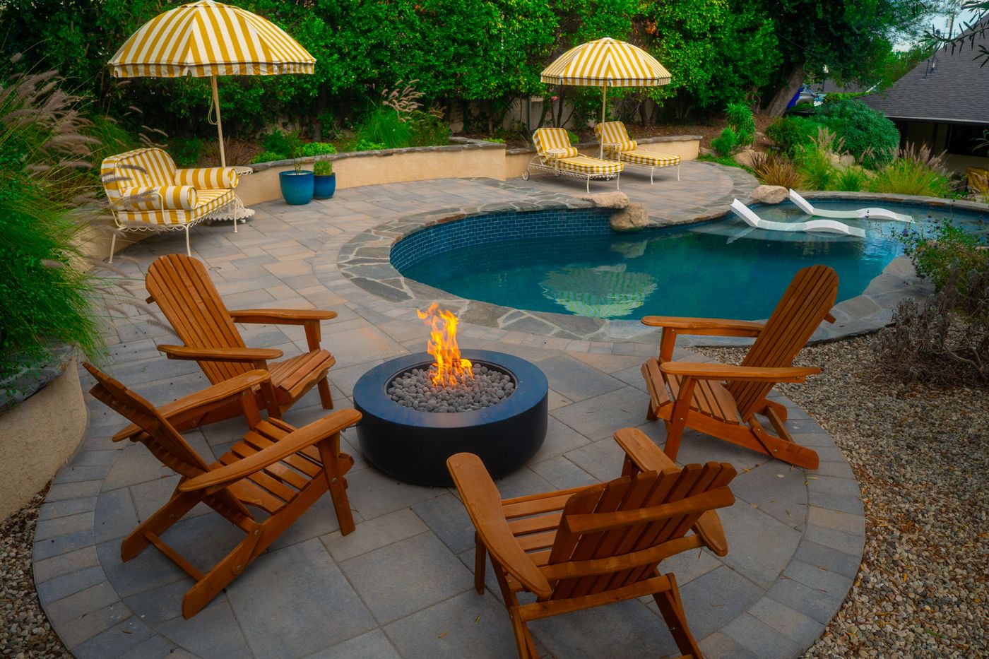 Backyard pool patio with round fire pit, Adirondack chairs, striped umbrellas, and layered greenery