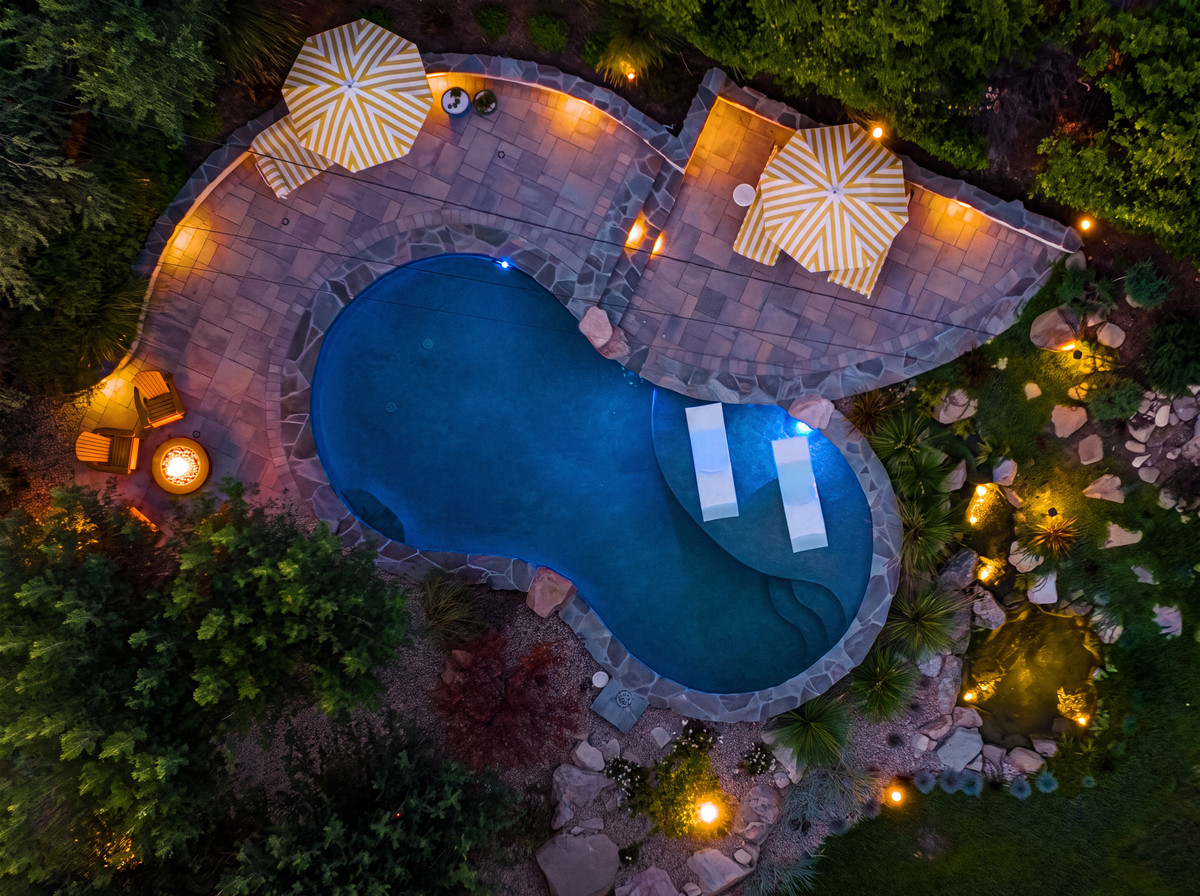 Aerial twilight view of a freeform pool and paver patio lit by landscape lighting, with uplit boulders, path lights, and a fire pit