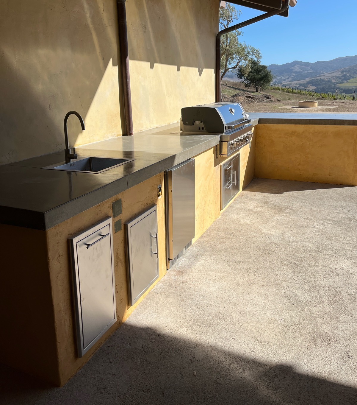 Completed outdoor kitchen with concrete counters, a stainless grill, sink, and refrigerator below vineyard hills