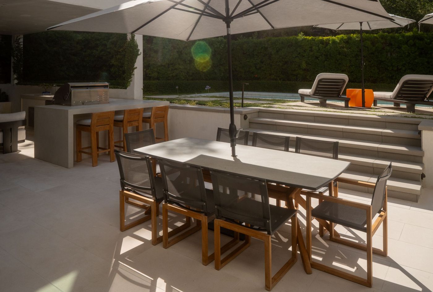 Modern patio dining space connected by steps to elevated poolside lounge chairs and landscaped greenery.