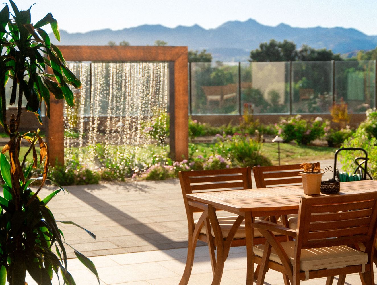 Patio dining table and chairs positioned beside a decorative water wall feature and landscaped courtyard garden with mountain backdrop.