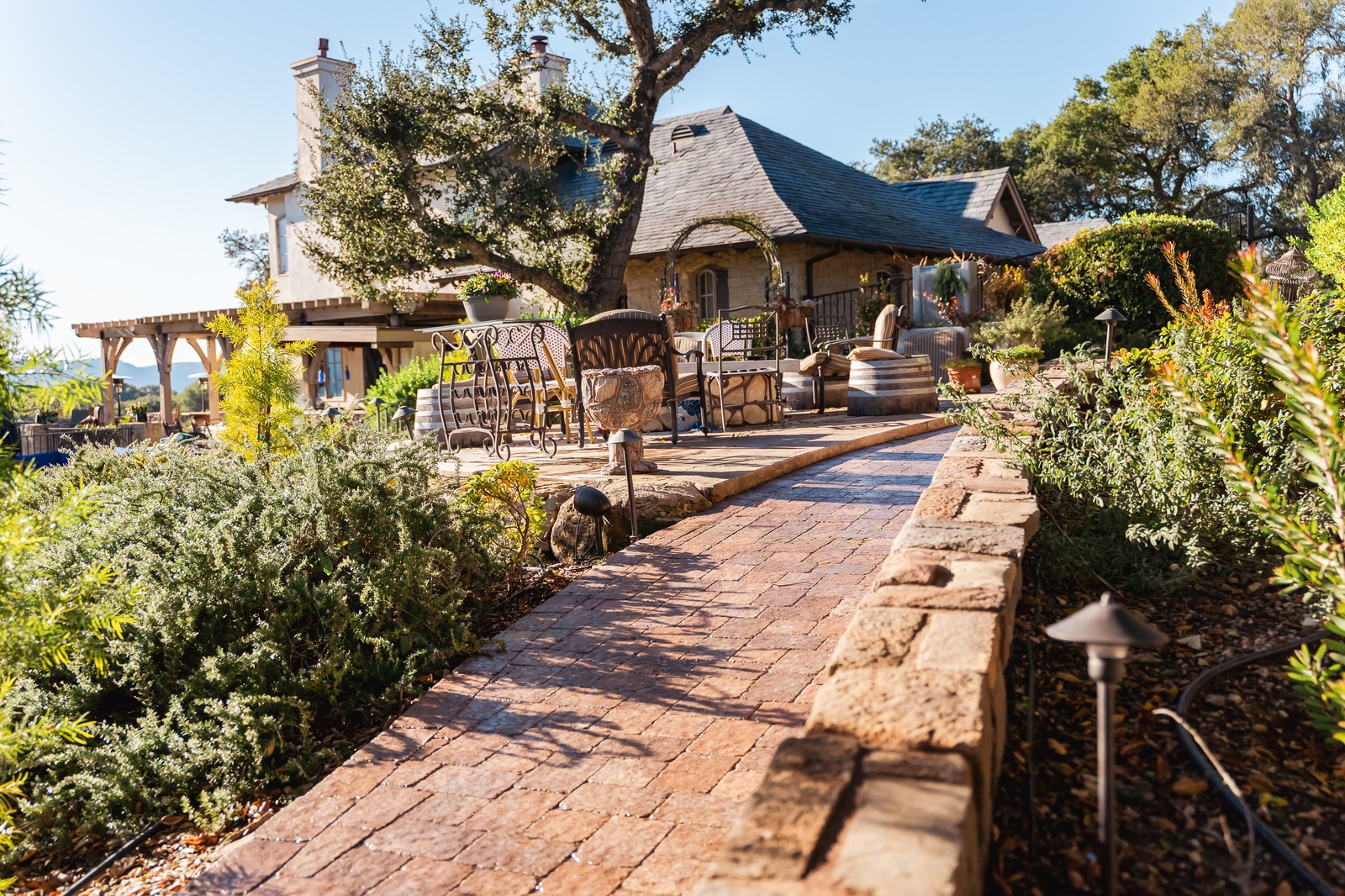 Brick paver walkway with a stone retaining wall leading to a stone house patio shaded by an oak