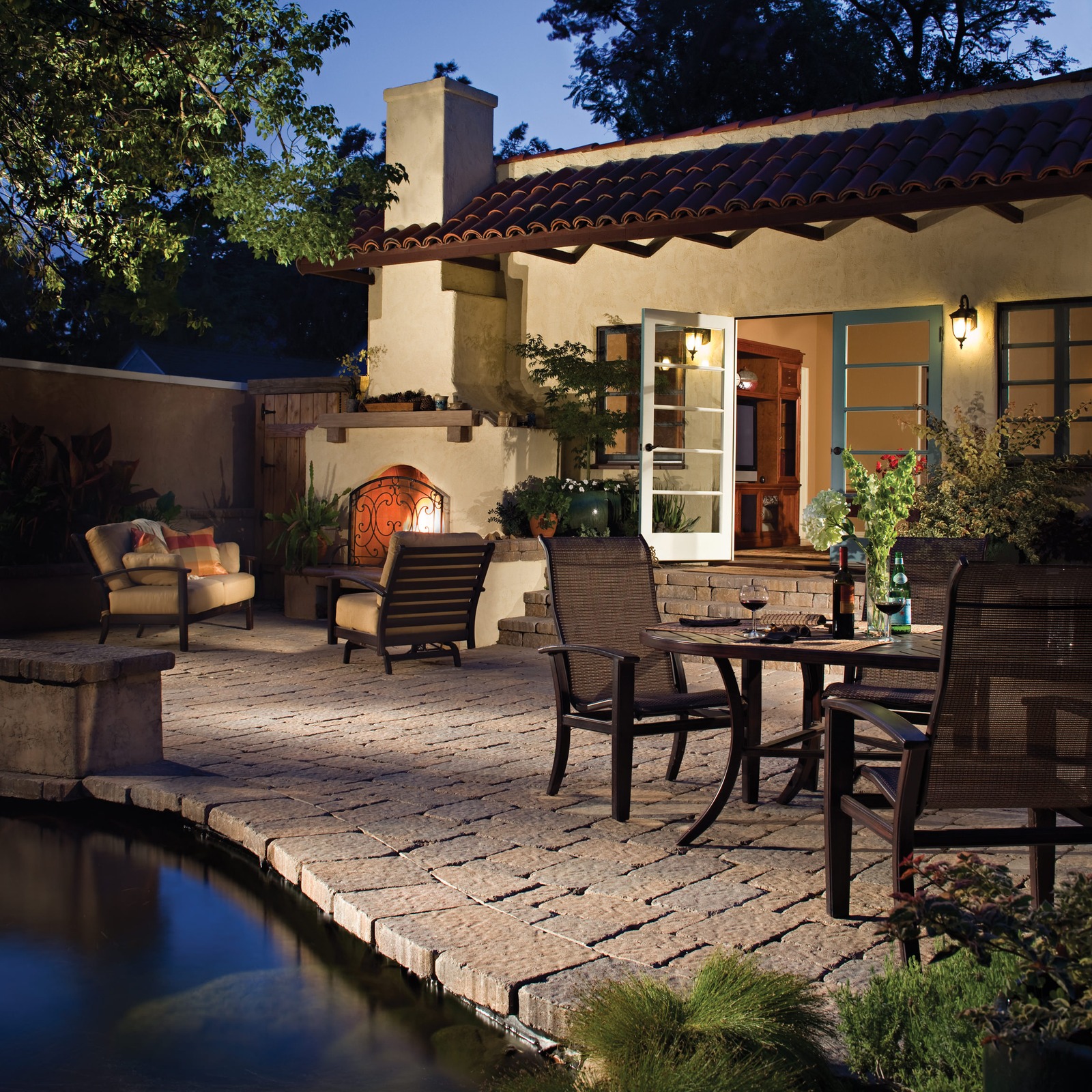 Twilight paver patio with a lit stucco fireplace, dining and lounge furniture, and open French doors