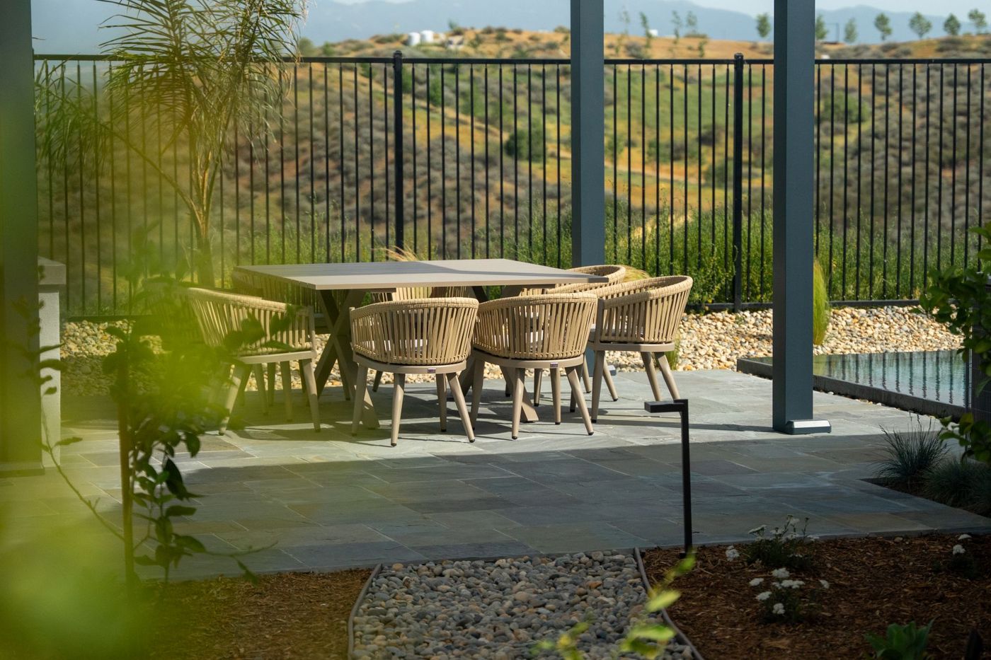 Outdoor dining table and woven chairs beneath sleek modern pergola next to long pool with hillside landscape backdrop.