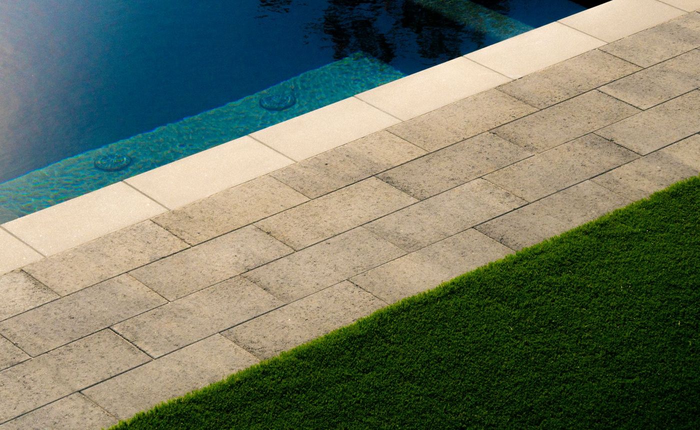 Close-up of swimming pool coping, stone pavers, and artificial turf in a modern landscaped backyard.