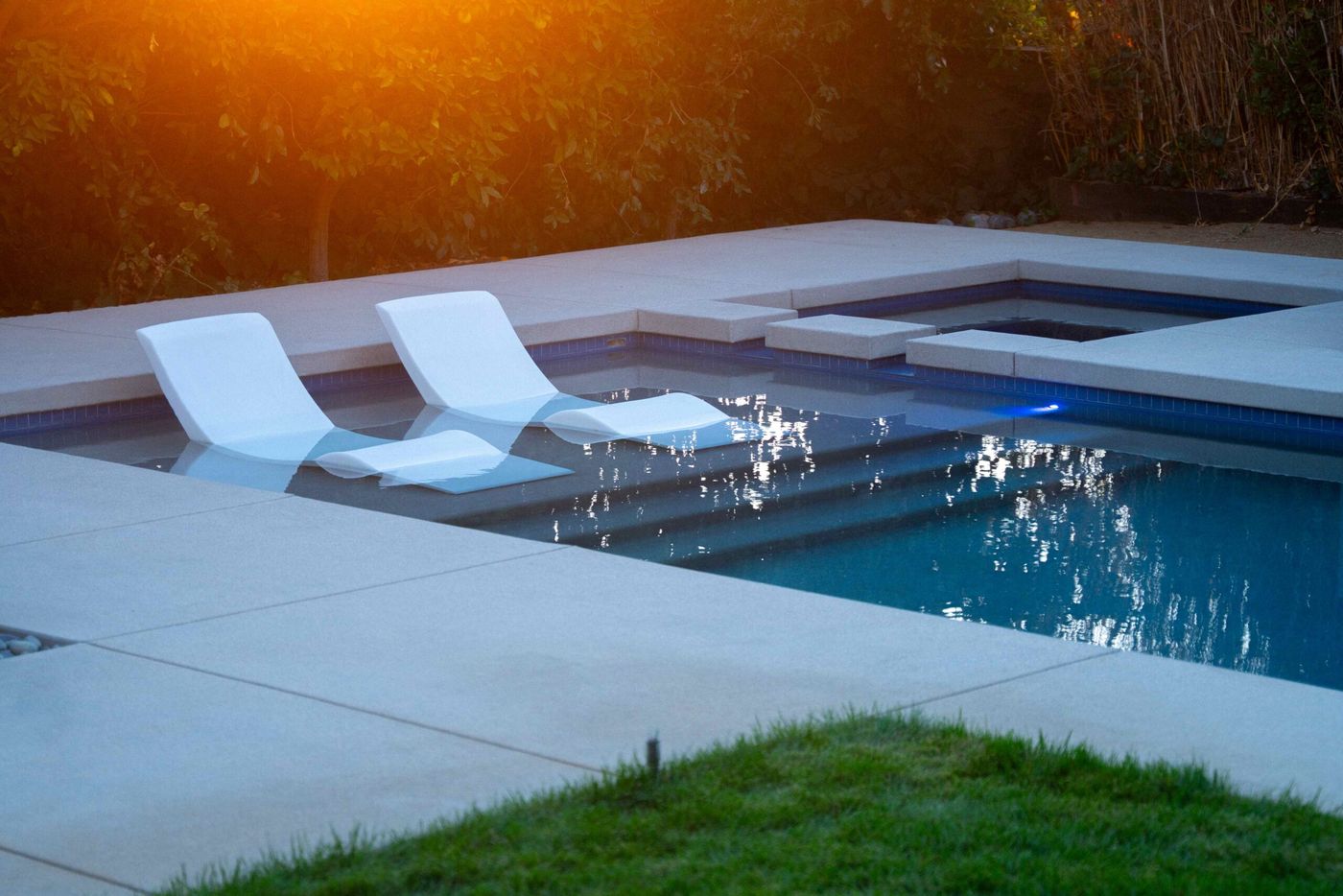 Swimming pool tanning shelf with white lounge chairs illuminated by warm sunset light in a modern backyard.