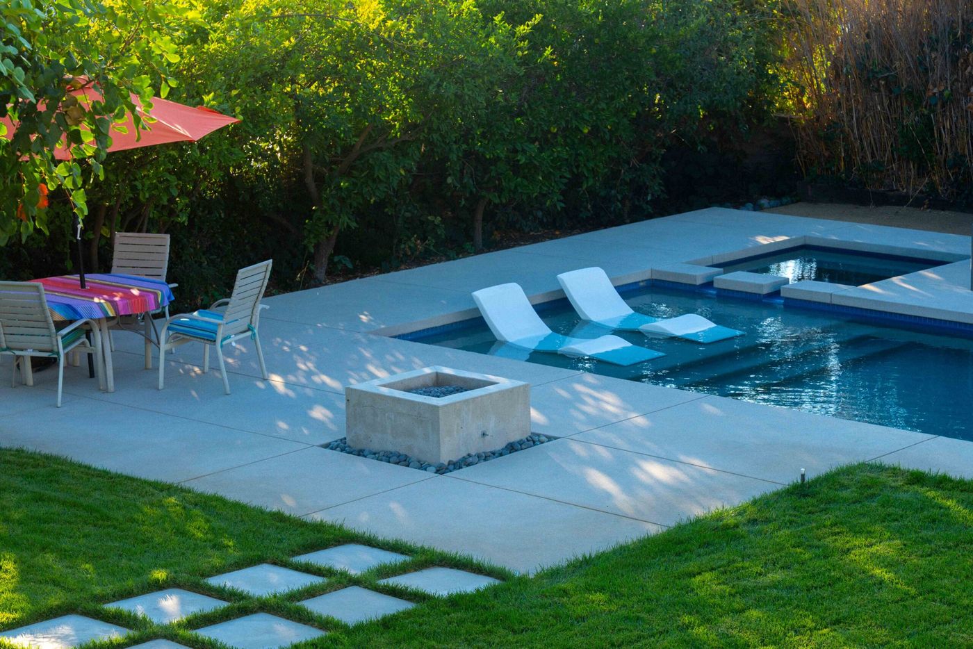 Contemporary backyard swimming pool with tanning ledge and white in-water lounge chairs surrounded by concrete decking and lawn.