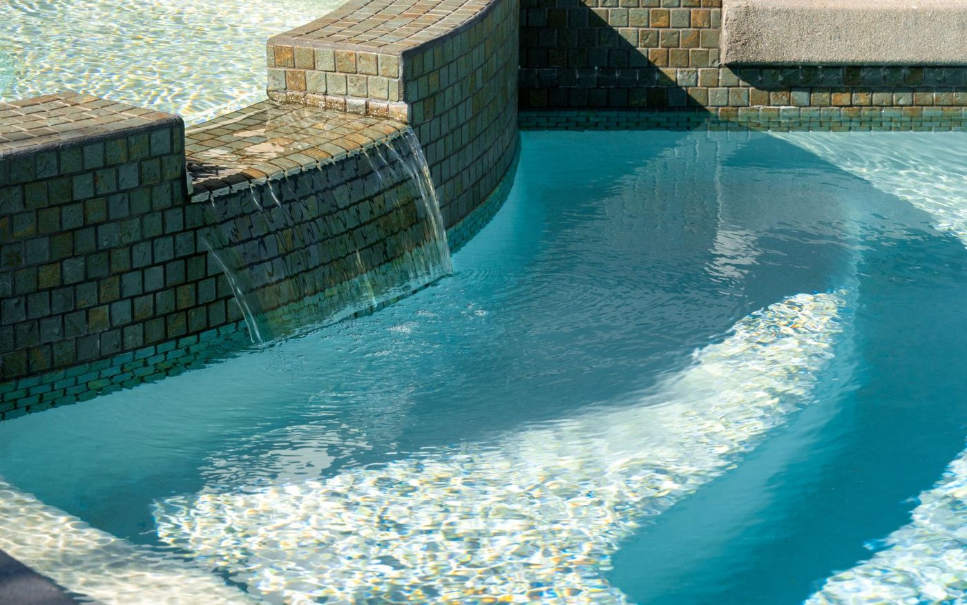 Close-up of tiled spa overflow waterfall flowing into clear blue swimming pool water.