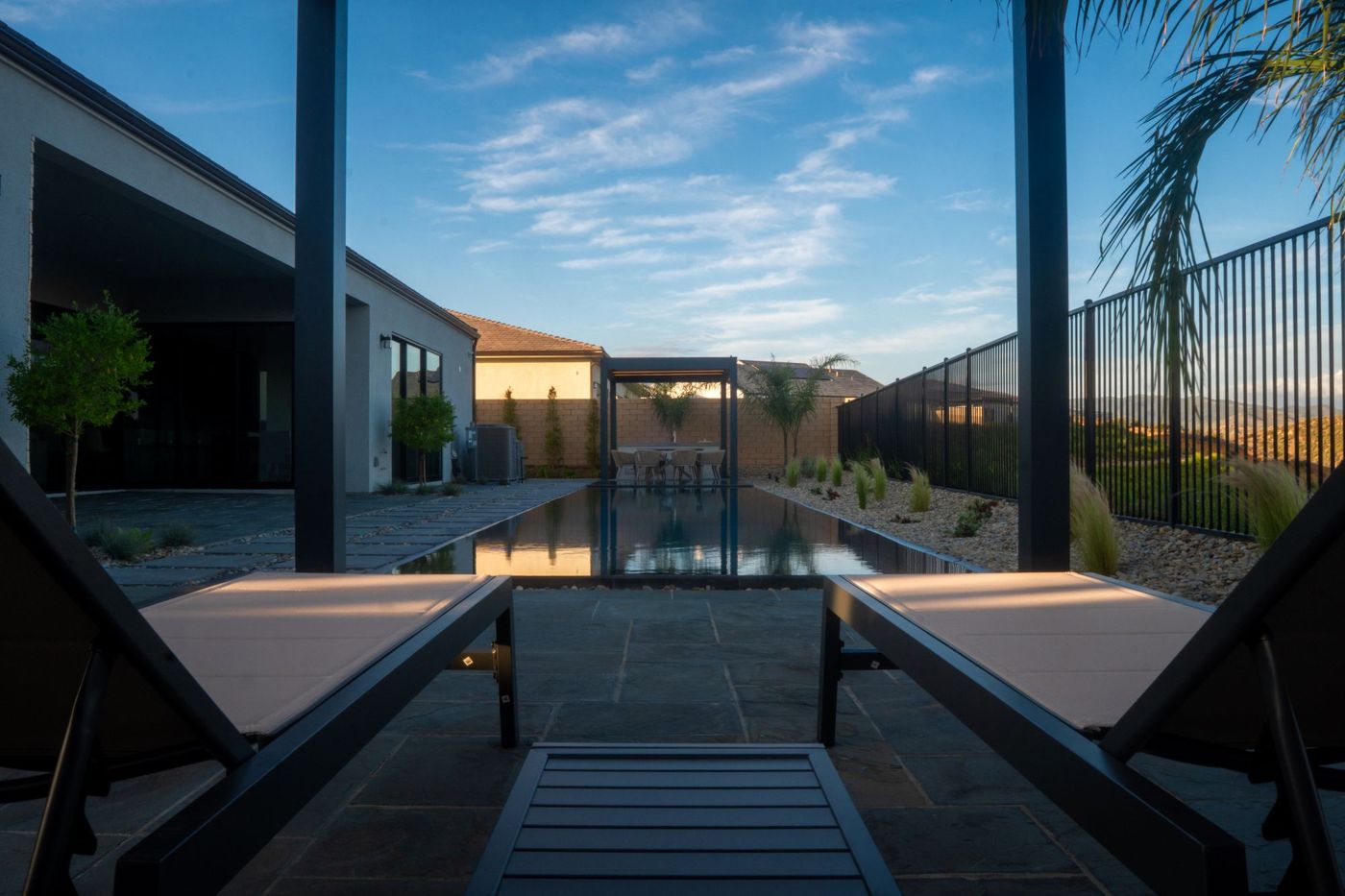 Two minimalist lounge chairs positioned under pergola looking toward long zero-edge reflecting pool and distant hillside landscape.