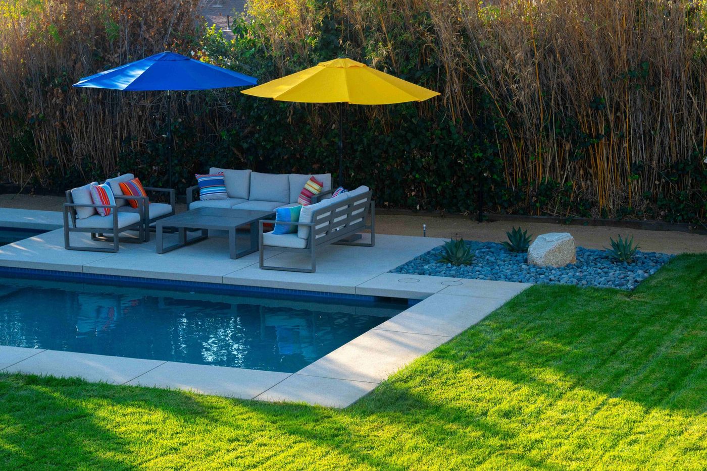 Modern poolside seating area with cushioned outdoor furniture, blue and yellow umbrellas, and concrete pool deck.